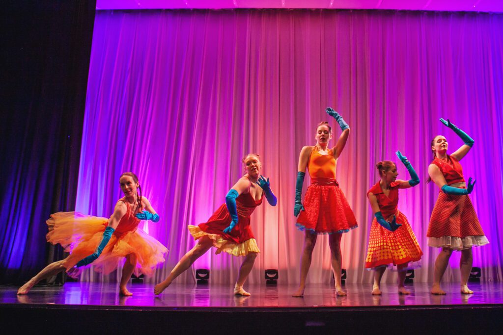 Five dancers perform on stage, each wearing vibrant red and yellow costumes with blue gloves. The background features a purple and pink illuminated curtain, adding a colorful atmosphere to the performance.
