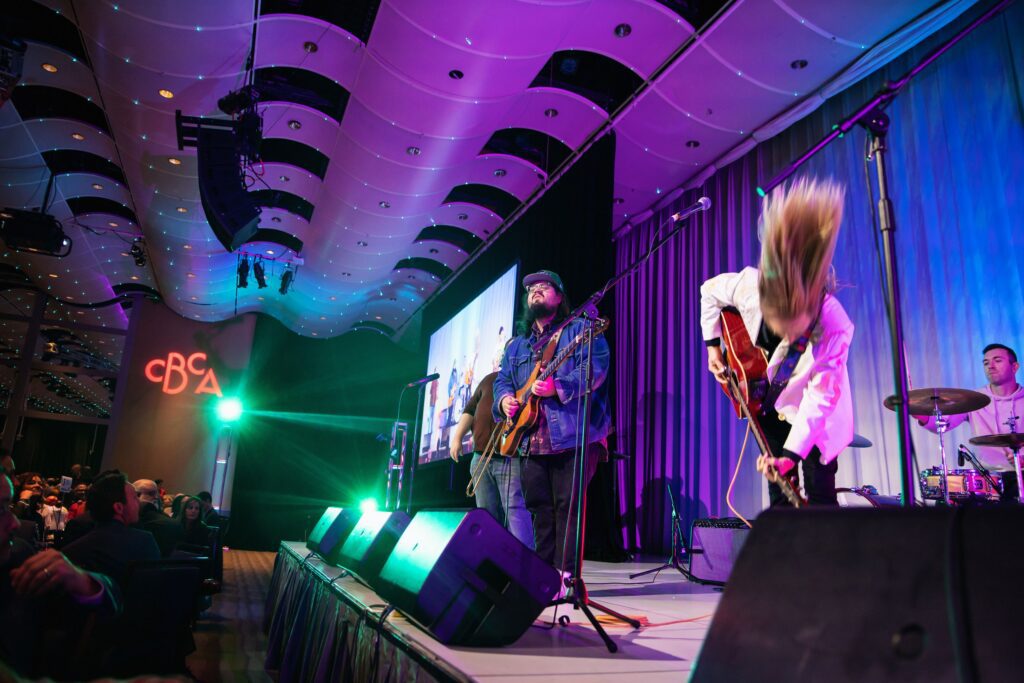 A band performing energetically on stage with vibrant lighting. The guitarist flips their hair, another musician plays bass, and a drummer is partially visible. The stage is set in a large venue with an audience watching, and a screen displays the performance.